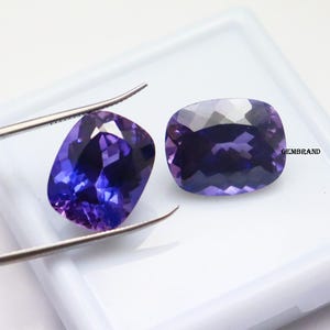 K&ouml;nnte beinhalten: Zwei kissenf&ouml;rmige Amethyst-Edelsteine, einer mit einer Pinzette gehalten, auf wei&szlig;em Hintergrund. Die Edelsteine sind tiefviolett mit einem leichten bl&auml;ulichen Schimmer.