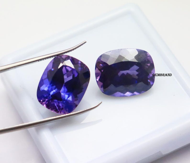 K&ouml;nnte beinhalten: Zwei kissenf&ouml;rmige Amethyst-Edelsteine, einer mit einer Pinzette gehalten, auf wei&szlig;em Hintergrund. Die Edelsteine sind tiefviolett mit einem leichten bl&auml;ulichen Schimmer.