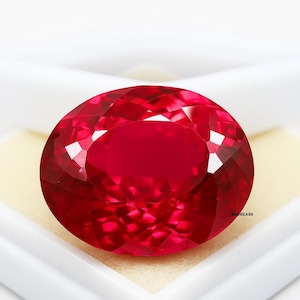 May include: A large, faceted, oval-shaped ruby gemstone. The gem is a deep, vibrant red color with multiple facets that reflect light. The ruby is set against a white background.