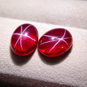 Pode incluir: Duas pedras preciosas ovais, vermelho escuro, com um padrão estelar de linhas brancas irradiando do centro. As pedras têm uma superfície polida e brilhante, e o efeito de estrela é proeminente. As pedras são provavelmente rubis.