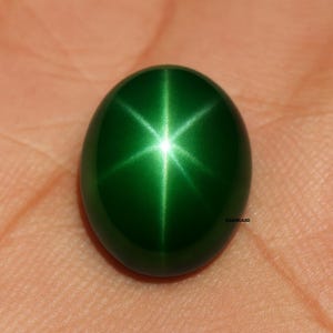 Könnte beinhalten: Ein ovaler, polierter grüner Edelstein mit einem sechsstrahligen Sterneffekt. Der Edelstein hat eine leuchtende smaragdgrüne Farbe und ist hochreflektierend. Der Edelstein befindet sich vor einem hautfarbenen Hintergrund.