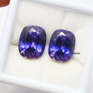 K&ouml;nnte beinhalten: Zwei Kissen-geschliffene Edelsteine, einer lila und einer blau, werden in einer wei&szlig;en Schachtel pr&auml;sentiert. Die Edelsteine sind facettiert und haben einen hohen Grad an Brillanz.