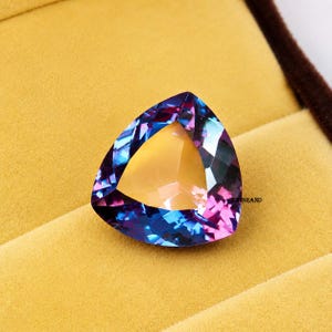May include: A triangular gemstone with a mix of blue, purple, and yellow hues. The gem is set against a yellow background, with a brown border visible in the upper right corner. The gem's facets reflect light, creating a sparkling effect.