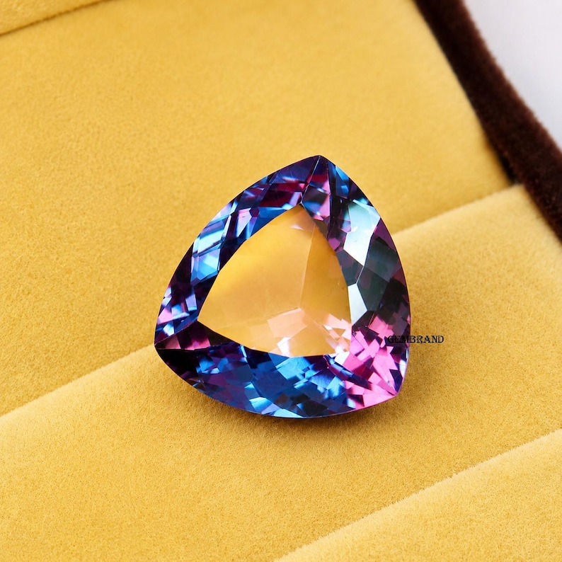 May include: A triangular gemstone with a mix of blue, purple, and yellow hues. The gem is set against a yellow background, with a brown border visible in the upper right corner. The gem's facets reflect light, creating a sparkling effect.