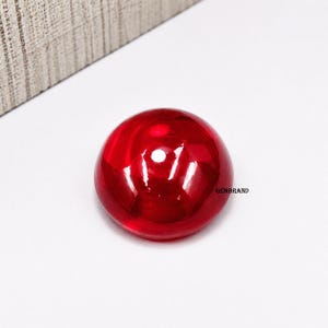 Op de afbeelding: Een gladde, cirkelvormige edelsteen in cabochon-stijl met een diepe, doorschijnende rode kleur en een hoogglanzend, reflecterend oppervlak. De steen heeft een gewelfde bovenkant en is voorzien van de zwarte tekst GEMBRAND op de zijkant.