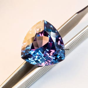 May include: A faceted, triangular gemstone with a color-shifting effect. The gem transitions from a golden hue to shades of blue and purple. Held by silver tweezers, the gem is displayed against a white background.