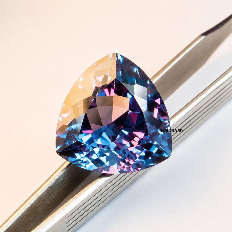 May include: A faceted, triangular gemstone with a color-shifting effect. The gem transitions from a golden hue to shades of blue and purple. Held by silver tweezers, the gem is displayed against a white background.