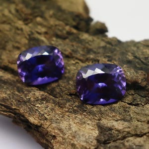 K&ouml;nnte beinhalten: Zwei violette Kissen-Schliff-Edelsteine auf einer rauen Holzoberfl&auml;che.