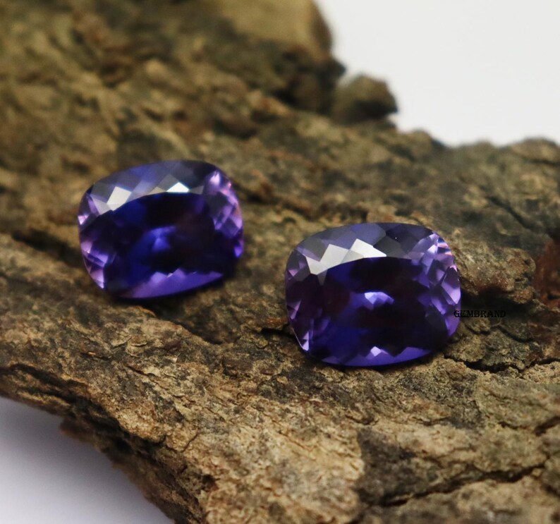 K&ouml;nnte beinhalten: Zwei violette Kissen-Schliff-Edelsteine auf einer rauen Holzoberfl&auml;che.
