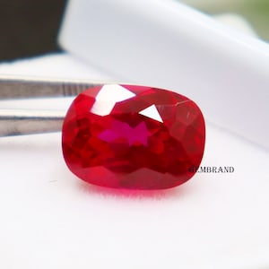 Puede incluir: Una gema ovalada de color rojo vibrante con un corte facetado. La gema está colocada sobre un fondo blanco.