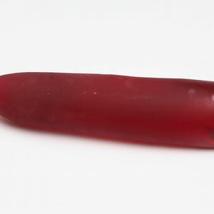 May include: A translucent, elongated, ruby-red gemstone with a pointed tip. The polished surface reflects light, showcasing the rich color and internal clarity of the mineral. The gemstone is displayed against a white background.