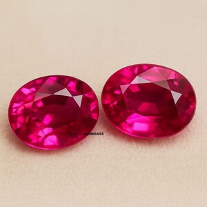 May include: Two oval-cut, vibrant red gemstones. The gems have multiple facets that reflect light, creating a sparkling effect. The stones are set against a neutral background, highlighting their rich color and clarity. The gemstones are likely rubies.