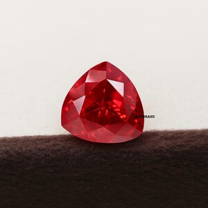 May include: A triangular, red gemstone with multiple facets. The gem is a deep, vibrant red colour, reflecting light. The gemstone is set against a neutral background, highlighting its brilliance and shape. Likely a ruby or similar precious stone.