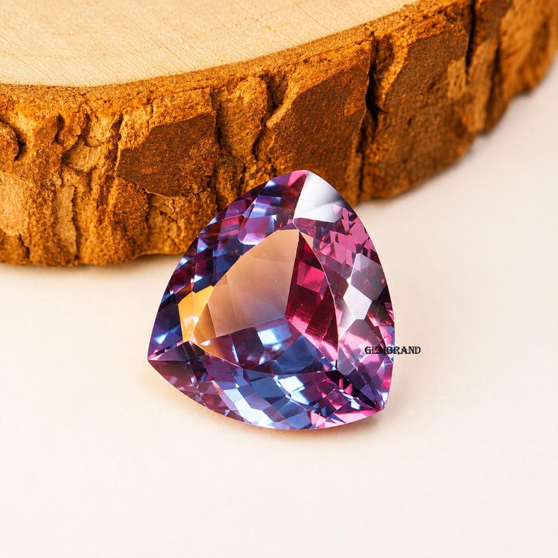 May include: A faceted, triangular gemstone with a blend of purple, blue, and pink hues. The gem is set against a light background, with a piece of textured wood in the upper portion of the image. The gem has a polished, reflective surface.