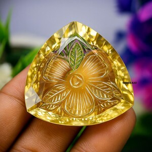 Puede incluir: Una piedra preciosa amarilla triangular tallada con un dise&ntilde;o floral. La piedra preciosa tiene un borde facetado y una talla detallada de una flor con m&uacute;ltiples p&eacute;talos y una hoja central. La piedra preciosa se sostiene en una mano.