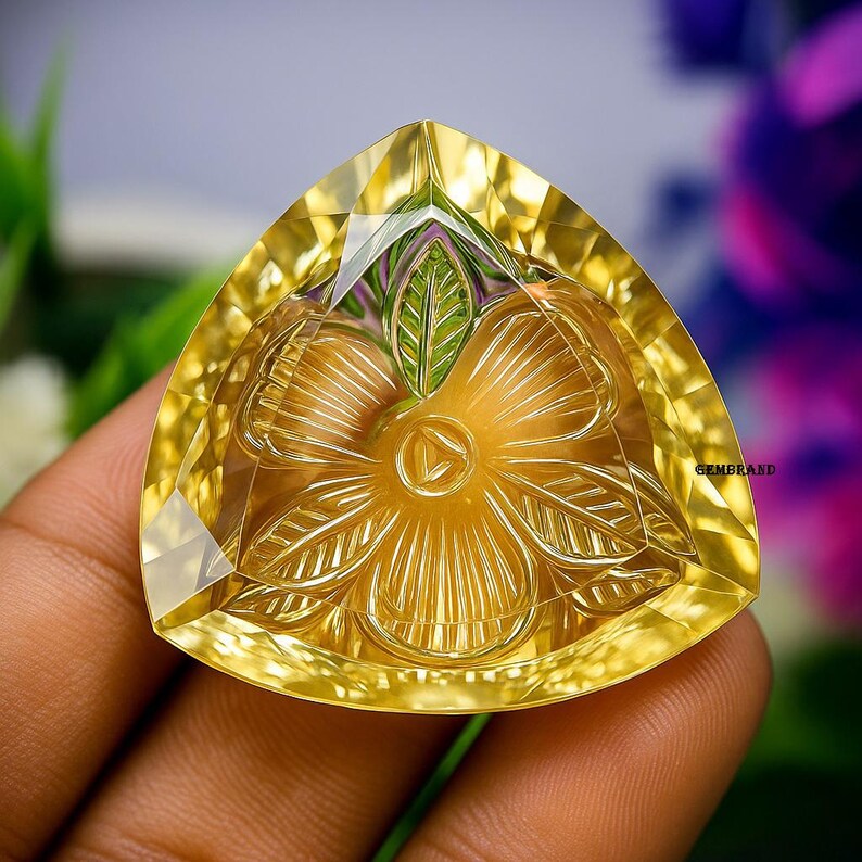 Puede incluir: Una piedra preciosa amarilla triangular tallada con un dise&ntilde;o floral. La piedra preciosa tiene un borde facetado y una talla detallada de una flor con m&uacute;ltiples p&eacute;talos y una hoja central. La piedra preciosa se sostiene en una mano.