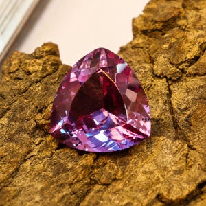 May include: A faceted, triangular gemstone with a blend of purple and pink hues. The gem is set against a textured, brown surface, showcasing its multiple facets and internal reflections. The gemstone is a striking example of fine jewelry.