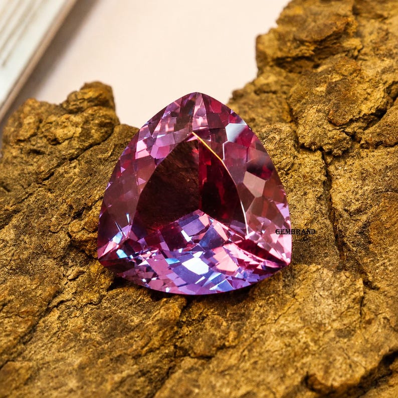 May include: A faceted, triangular gemstone with a blend of purple and pink hues. The gem is set against a textured, brown surface, showcasing its multiple facets and internal reflections. The gemstone is a striking example of fine jewelry.