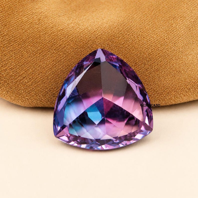 May include: A faceted, triangular gemstone with a gradient of colors, including purple, blue, and pink. The gem is cut to reflect light, creating a sparkling effect. The gemstone is set against a neutral background.