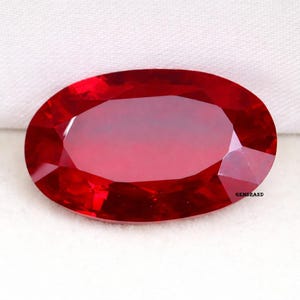 Pode incluir: Um rubi de corte oval, com uma cor vermelha intensa e saturada. A gema apresenta múltiplas facetas que refletem a luz. O rubi é exibido sobre um fundo branco, realçando sua cor e brilho. A imagem é um close-up que destaca os detalhes da pedra.