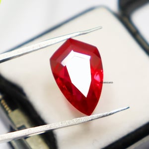 May include: A deep red pear-shaped gemstone held by tweezers. The gemstone is faceted and has a high polish. The background is a white surface with the text "GEMBRAND".