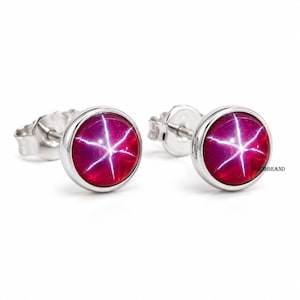 May include: A pair of stud earrings featuring round, deep red cabochon stones with a distinct six-rayed star effect. The stones are set in polished silver-toned metal settings with standard post backs. The text GEMBRAND is visible on the right.