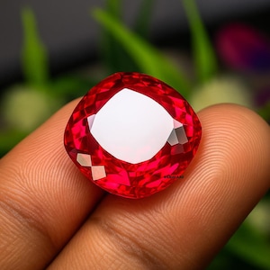 Może przedstawiać: Fasetowany, szlifowany w kształcie poduszki, czerwony kamień szlachetny z jasnym, białym środkiem. Kamień trzymany jest między palcami, ukazując jego wspaniały szlif i żywy kolor. Fasetki kamienia odbijają światło.
