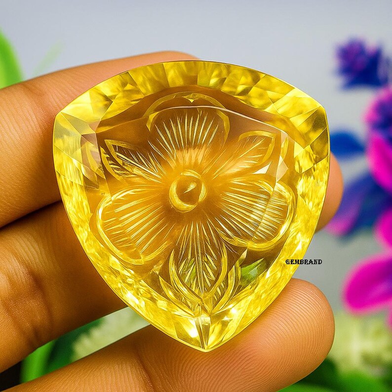 Puede incluir: Una gema triangular amarilla con un dise&ntilde;o floral tallado. La gema se sostiene en una mano y el fondo est&aacute; borroso. La gema tiene un borde facetado y un tallado floral detallado en el centro. El texto "GEMBRAND" es visible en la parte inferior derecha.