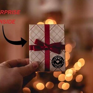 Puede incluir: Una peque&ntilde;a caja de regalo con un patr&oacute;n de cuadros blancos y beige, una cinta roja y un lazo. La caja tiene un logotipo circular con las palabras "GEMS BRAND". Las palabras "SURPRISE INSIDE" est&aacute;n encima de la caja.
