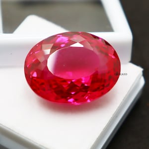 May include: A bright red, oval-cut gemstone, presented in a white plastic display case. The gem's surface is highly reflective, highlighting its facets. The text "EMIRAND" is visible on the side of the case.