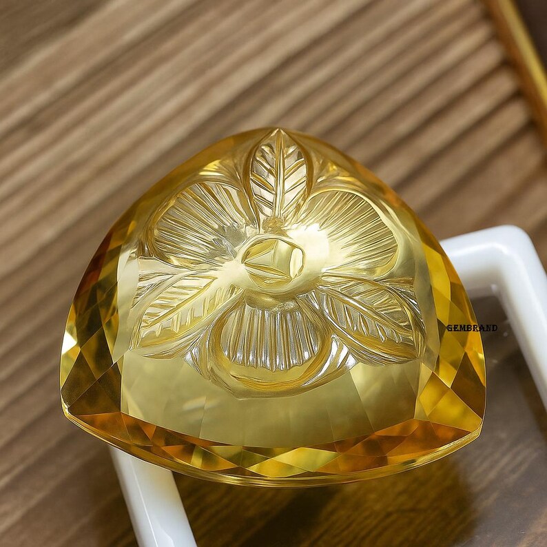 Puede incluir: Una gema amarilla facetada, de forma triangular, con intrincadas tallas florales. La gema es de un color amarillo vibrante y se encuentra sobre un fondo borroso. Es probable que la gema sea un art&iacute;culo decorativo o un componente de joyer&iacute;a.