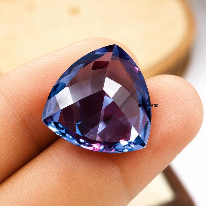 May include: A faceted, triangular gemstone with a color gradient from deep purple to blue. The gem is held in a hand, showcasing its polished surfaces and internal reflections. The gem's facets catch the light, creating a sparkling effect.