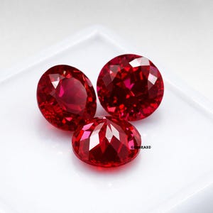 May include: Three faceted, oval-cut, deep red gemstones. The gems are displayed on a white surface, showcasing their brilliant, reflective surfaces. The gemstones are likely rubies, known for their rich color and luster.
