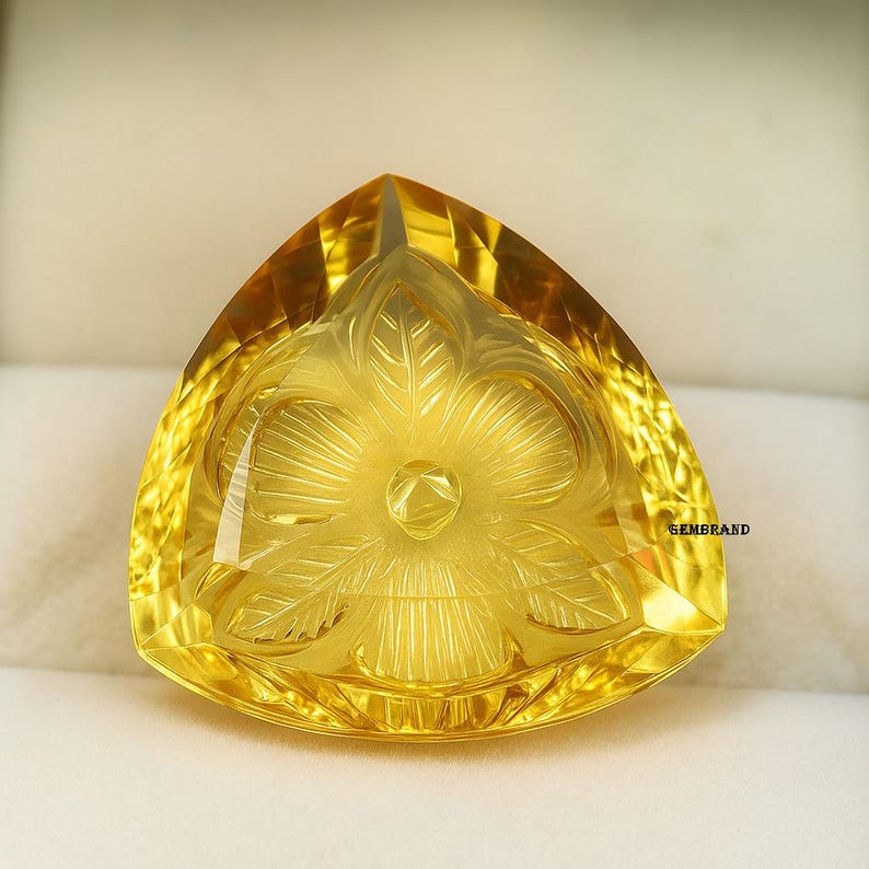 Puede incluir: Una gema triangular amarilla facetada con un dise&ntilde;o floral tallado. La superficie pulida refleja la luz. Es probable que sea un elemento decorativo o parte de una joya.