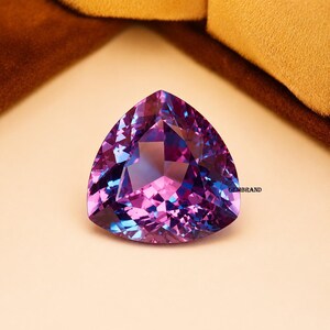 May include: A large, triangular gemstone with facets, displaying a blend of purple, blue, and pink hues. The gem has a polished surface and is set against a neutral background. The word "GEMBRAND" is visible on the right side of the gem.