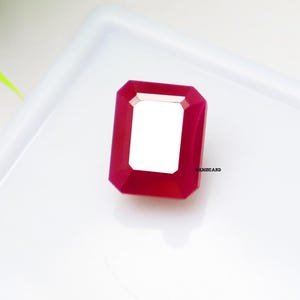 May include: A faceted, rectangular-cut ruby gemstone with a bright red hue. The gem has a white, rectangular center and is set against a white background. The gemstone is a vibrant example of fine jewelry.