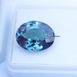 May include: An oval-shaped gemstone with a faceted surface. The gem displays a blend of blue and teal hues, with darker shades towards the edges. The gem is set against a white background. The text "GEMBRAND" is visible on the right side.