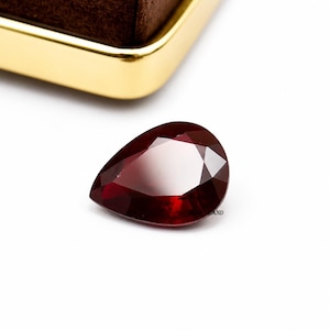 May include: A pear-shaped, deep red faceted gemstone with a polished surface. The stone features multiple triangular and kite-shaped facets that reflect light, creating a brilliant appearance. The gem is positioned on a plain white surface.
