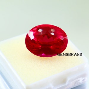 May include: A faceted, oval-shaped, vibrant red gemstone. The gem is displayed on a white surface inside a clear plastic container. The text "GEMSBRAND" is visible on the container. The gemstone's color is a deep, saturated red.