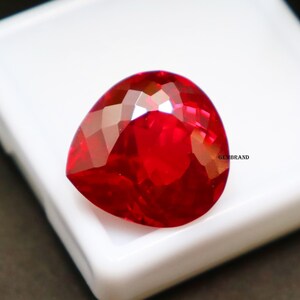 May include: A deep red gemstone with a heart shape cut. The gemstone is on a white surface.