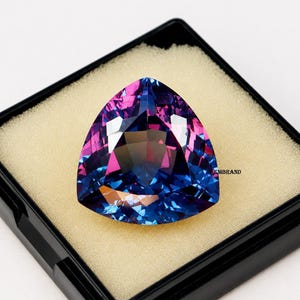 May include: A large, triangular gemstone with a vibrant color gradient of pink, purple, and blue. The gem is faceted, displaying multiple reflective surfaces. It is set in a black display case with a beige interior.