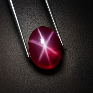 Puede incluir: Una gema de rubí pulida, de forma ovalada, con un efecto de estrella de seis rayos, sujeta por unas pinzas plateadas. El rubí es de color rojo intenso con un patrón de estrella blanco brillante. Fondo oscuro y texturizado.