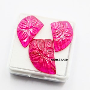 May include: Three pink carved gemstone cabochons in a white box. The cabochons are shaped like half circles and have a floral design carved into them. The box has the text "GEMSBBRAND" printed on it.