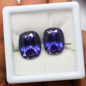K&ouml;nnte beinhalten: Zwei violette Kissen-Schliff-Edelsteine in einer wei&szlig;en Schachtel. Die Edelsteine sind etwa 10 mm x 8 mm gro&szlig;.