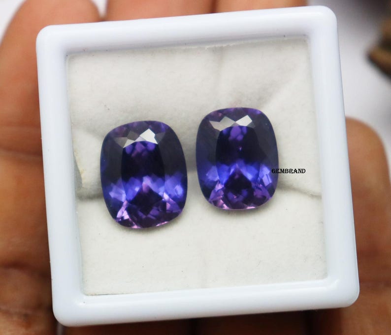 K&ouml;nnte beinhalten: Zwei violette Kissen-Schliff-Edelsteine in einer wei&szlig;en Schachtel. Die Edelsteine sind etwa 10 mm x 8 mm gro&szlig;.