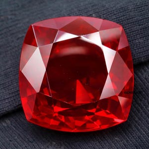 Könnte beinhalten: Ein facettierter, kissenförmiger Edelstein in einem leuchtenden, tiefroten Farbton. Die Oberfläche des Edelsteins reflektiert das Licht und zeigt seine komplizierten Schnitte und Winkel. Der Edelstein ist vor einem dunklen Hintergrund platziert.
