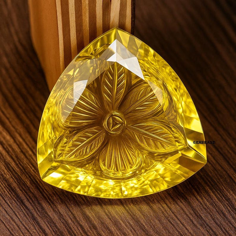 Puede incluir: Una gema amarilla triangular y facetada con un dise&ntilde;o floral tallado en su superficie. La gema se encuentra sobre un fondo de madera, mostrando su claridad y detalles intrincados. La gema es de un color amarillo vibrante.