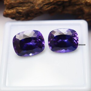 K&ouml;nnte beinhalten: Zwei Amethyst-Edelsteine im Kissen-Schliff, jeder mit einem tiefen Purpurton, werden auf einer wei&szlig;en Oberfl&auml;che pr&auml;sentiert. Die Edelsteine sind etwa 10 mm x 15 mm gro&szlig;.