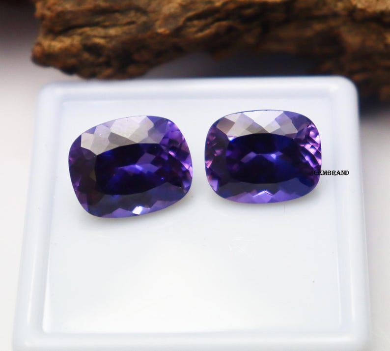 K&ouml;nnte beinhalten: Zwei Amethyst-Edelsteine im Kissen-Schliff, jeder mit einem tiefen Purpurton, werden auf einer wei&szlig;en Oberfl&auml;che pr&auml;sentiert. Die Edelsteine sind etwa 10 mm x 15 mm gro&szlig;.