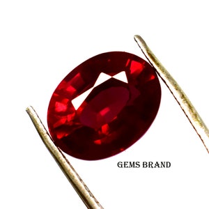 Puede incluir: Una piedra preciosa ovalada de color rojo intenso sostenida en una pinza. La piedra preciosa está facetada y tiene un alto brillo. El texto "GEMS BRAND" es visible en la esquina inferior derecha de la imagen.